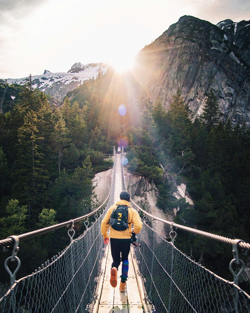 A hiker crossing a suspension bridge toward the sunrise between mountain peaks